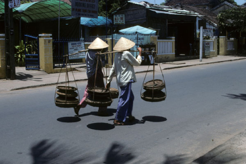 Two women carry trays suspended from shoulder poles in street at Hoi An